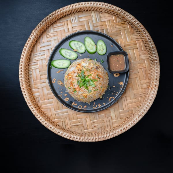 Laos fried rice with sliced cucumbers and dipping sauce on a black plate, on a woven tray.