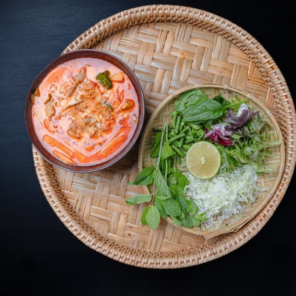 Laos red curry soup with noodles and meat, served with fresh herbs and lime on a woven tray.