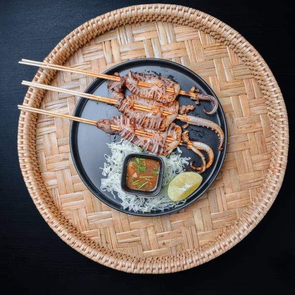 Grilled squid skewers with dipping sauce, shredded cabbage, and lime on a black plate.