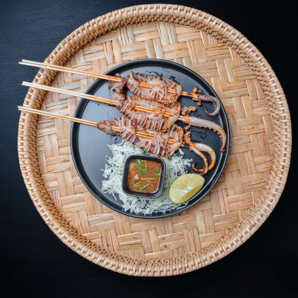 Grilled squid skewers with dipping sauce, shredded cabbage, and lime on a black plate.